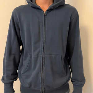 Blå hoodie med dragkedja från Uniqlo - Mörkblå hoodie från Uniqlo i mjuk bomullsmix. Tröjan har hel dragkedja, två stora fickor framtill och en skön huva. Perfekt för chill dagar eller när du vill ha en enkel och clean look.