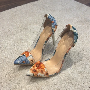 Blommiga pumps med transparent sida - Snygga pumps med spetsig tå och hög klack. Skorna har blommigt mönster i orange, blått och vitt samt transparenta sidor i plast som ger en modern vibe. Perfekta för dig som vill sticka ut med färg och cool design.
