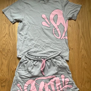 Syna world tracksuit - Syna world tracksuit, tröja + shorts. Snygga grå shorts med stort rosa grafiskt tryck framtill och matchande rosa snörning i midjan. Mjukt material och avslappnad passform, perfekta för sommardagar eller chill hemma. Elastisk midja för extra komfort och cool streetkänsla.