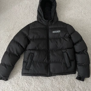 Svart pufferjacka från Hoodrich - Säljer en svart pufferjacka från Hoodrich med stor logga på ryggen och mindre logga på bröstet. Jackan har huva, dragkedja och två snedställda fickor framtill. Perfekt för kalla dagar och riktigt street i stilen.