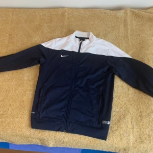 Nike Dri-Fit vindjacka blå/vit L - 🚨Pris kan diskuteras 🚨Nike Dri-Fit vindjacka i marinblått och vitt med dragkedja framtill och två fickor. Jackan har hög krage, lång ärm och klassisk Nike-logga på bröstet och ryggen. Perfekt för sport eller chill dagar. Materialet är lätt och andas.