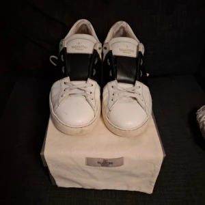 Valentino Garavani vita sneakers - Snygga vita sneakers från Valentino Garavani med bred svart läderdetalj över vristen och klassisk snörning. Skorna har rund tå, platt sula och diskret logga på plösen. Kommer med originalpåse och box. Perfekta för dig som gillar stilrena designer. OBS boxen är inte valentinos orginal, tveka inte på att ställa frågor, skicka gärna prisförslag (jag är öppen för det mesta) och skriv om ni vill ha fler bilder. Skorna är i storlek 41