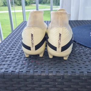 Nike Mercurial Air Zoom fotbollsskor vita - Snygga Nike Mercurial Air Zoom fotbollsskor i vitt med svarta detaljer och hög krage. Skorna har en strömlinjeformad design, stora svarta swoosh-loggor och texten 'AIR' på sidan. Perfekta för dig som vill sticka ut på planen.