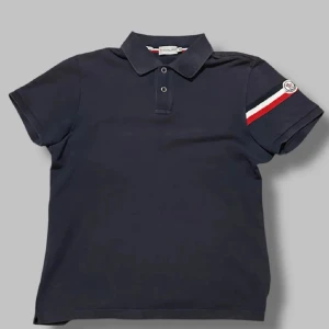 Moncler Piké - Säljer en riktigt snygg mörkblå pikétröja från Moncler med korta ärmar och en klassisk krage. Självklart äkta men en äldre modell som har en broderad Moncler-logga på ärmen. Vid minsta fråga eller fundering, tveka inte på att höra av dig🤝