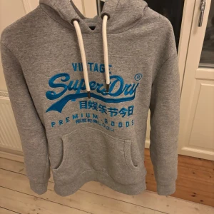 Superdry hoodie - Säljer en grå hoodie från Superdry med blå logga och text framtill. Tröjan har huva med vita snören och en stor magficka. Perfekt för en avslappnad stil och enkel att matcha med det mesta. Pytte pytte små röda märken
