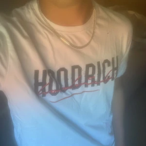 Vit HOODRICH t-shirt med tryck - Vit t-shirt från HOODRICH med svart och rött tryck framtill. Klassisk rund hals och korta ärmar. Snygg detalj med broderad röd text över loggan och patch nertill. Perfekt för en streetwear-look. Det finns några hål på den men man tänker inte på det så mycket. Tveka inte på att höra av dig!