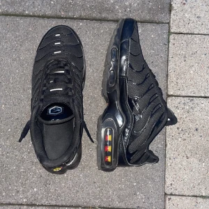 Nike Air Max Plus Tn i helsvart - Helt nya Nike Air Max Plus Tn i helsvart med mesh och syntetpaneler, ikoniska vågformade detaljer och synlig Air Max-dämpning i sulan. Skorna har snörning och Tn Air-logga på hälen och sulan. Perfekta för dig som gillar streetwear och sportig stil.