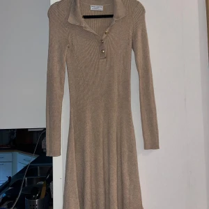 Beige ribbad kortklänning Lindex  - Snygg beige kortklänning från Lindex med ribbad struktur, krage och tre dekorativa knappar framtill. Klänningen har lång ärm och är figurnära upptill med lite vidare kjol. Perfekt för dig som gillar stilren och enkel design.