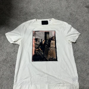 Vit Limitato t-shirt med tryck - Vit t-shirt från Limitato med ett stort, färgstarkt fototryck på framsidan som visar en person på en balkong med stadsmiljö i bakgrunden. T-shirten har rund hals och korta ärmar, tillverkad i mjuk bomull. Limitato-logga i vitt på ryggen. Nypris 3000kr, använd sparsamt och ett fåtal gånger. Max 3-4 gånger.