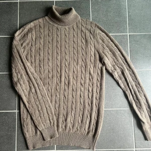 Beige polotröja från John Henric - Säljer en beige kabelstickad polotröja från John Henric. Tröjan har hög polokrage, lång ärm och klassiskt flätmönster. Perfekt för lager-på-lager och chill höststil. Materialet är mjukt och stickat för extra komfort.