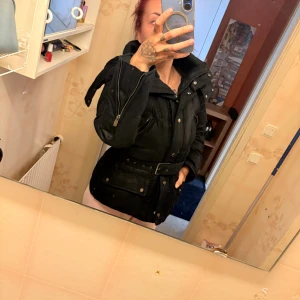 Svart parkajacka med bälte och dragkedja - Säljer en svart parkajacka med bälte i midjan och flera fickor framtill. Jackan har hög krage, långa ärmar med dragkedjedetaljer och knappar. Perfekt för kyligare dagar och riktigt snygg passform.