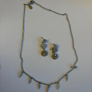 Glittrigt silverset halsband & örhängen - Snyggt silverset med halsband och matchande örhängen. Halsbandet har små hängande blixtar och örhängena är dubbla med gnistrande stenar. Perfekt för dig som vill sticka ut med lite extra bling och coola detaljer. Ej, använda!