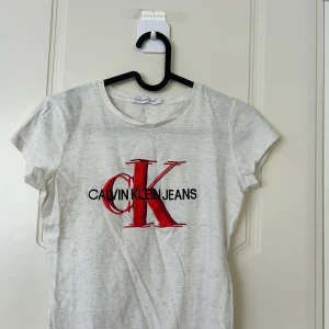 Vit t-shirt Calvin Klein Jeans - Vit t-shirt från Calvin Klein Jeans med stort CK-logotryck i rött och svart på bröstet. Rund hals och korta ärmar. T-shirten är gjord i mjuk bomull och har en clean, klassisk look som passar perfekt till jeans eller shorts.