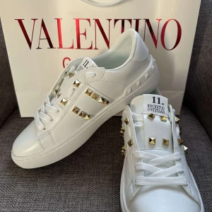 Valentino Rockstud vita sneakers - Snygga vita Valentino Rockstud sneakers i skinn med ikoniska guldiga nitar på sidorna och tungan. Klassisk rund tå, platt sula och vita snören. Perfekta för dig som vill sticka ut med en lyxig och edgy vibe.
