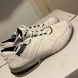 Vita sneakers från Puma med svarta detaljer - Snygga vita sneakers från Puma med svarta detaljer och logga på sidan, tungan och hälen. Skorna har snörning, perforerade partier för ventilation och en robust sula med silvriga inslag. Perfekta för dig som gillar sportig och clean stil.