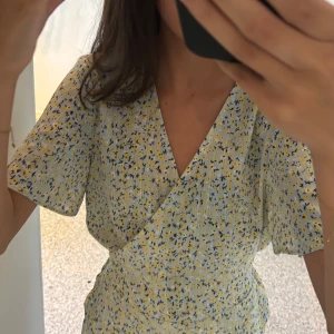 Blommig omlottblus från Vila - Supersöt kort omlottblus från Vila i ljusblått med små gula och svarta blommor. Blusen har v-ringning, korta ärmar och knyt i sidan. Perfekt för sommarens alla tillfällen, riktigt fräsch och lätt i materialet.