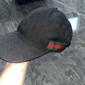 Svart Gucci keps med monogram - Säljer en svart keps från Gucci med diskret GG-monogram över hela kepsen och klassiska gröna och röda ränder på sidorna. Justerbar rem baktill i skinn och tillverkad i Italien. Perfekt för dig som vill ha en lyxig och stilren accessoar.