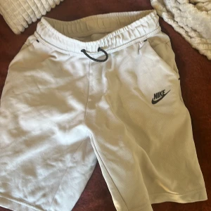 Vita Nike shorts med snörning XL 170 - Snygga vita shorts från Nike i storlek XL. De har elastisk midja med svart snörning, sidofickor och en bakficka med dragkedja. Perfekta för träning eller chill dagar. Standard fit och tillverkade i mjukt bomullsmaterial.