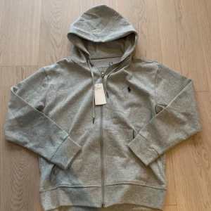 Grå hoodie från Polo Ralph Lauren - Snygg grå hoodie från Polo Ralph Lauren med dragkedja och huva. Klassisk broderad logga på bröstet och praktiska fickor framtill. Tillverkad i mjuk bomullsmix, perfekt för chill dagar eller när du vill ha en clean look., OBS 🐼helt ny