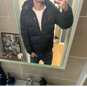 Svart pufferjacka Polo Ralph Lauren - Säljer en svart pufferjacka från Polo Ralph Lauren med huva och dragkedja. Jackan har flera fickor med dragkedja och en klassisk quiltad look. Perfekt för kalla dagar och riktigt snygg till jeans eller sweatpants.