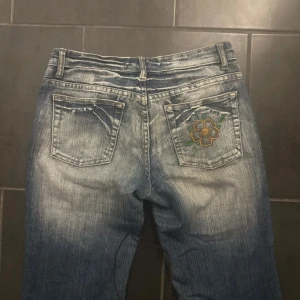 Blå broderade jeans med blommor - Snygga blå jeans med slitningar och broderade blommor i grönt och gult på benen och bakfickan. Klassisk femficksmodell med normal passform och bootcut. Jeansen har bälteshällor och dragkedjegylf. Perfekta för dig som vill sticka ut lite extra!