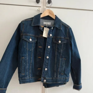 Klassisk blå jeansjacka från Monki - Snygg blå jeansjacka från Monki med silverfärgade knappar och klassisk krage. Jackan har två bröstfickor med lock och knappar samt två sidofickor. Perfekt för lager-på-lager och enkel att styla till allt.