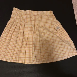 Rutig gul kortkjol från Urban Outfitters - Superfin plisserad kortkjol från Urban Outfitters i ett rutigt mönster med gula och vita toner. Kjolen har en liten ficka med UO-76 patch och dragkedja i sidan. Perfekt för dig som gillar en söt och trendig stil.