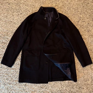 loro piana spagna coat - Snygg mörkblå rock med stilren ståkrage och stora fickor framtill. Jackan har knäppning med knappar och diskreta detaljer i mörkgrått längs kragen. Fodret är mörkt och insidan av fickorna har en brun accent. Perfekt för dig som gillar klassisk men modern stil.