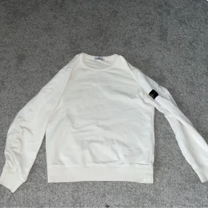 Vit Stone Island Junior sweatshirt - Vit sweatshirt från Stone Island Junior med klassisk logga på ärmen. Tröjan har rund hals, ribbade muddar och är tillverkad i mjuk bomull. Perfekt för dig som gillar stilrena och ikoniska plagg med streetkänsla. Kan gå ner i pris vid snabbt köp. Passar lika bra som 158,164. Kanske byte beroende på vad du erbjuder. 