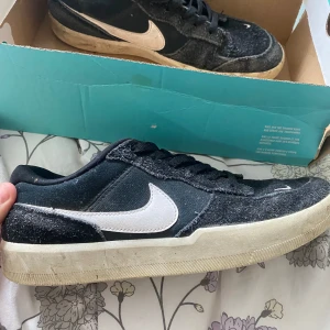 Svarta Nike SB  - Svarta Nike SB sneakers med vit swoosh och klassisk låg profil. Ovandelen är i ruggad mocka och textil, med svarta skosnören och vit sula. Perfekta för skate eller streetstyle. Snygg kontrast mellan svart och vitt, och diskret Nike-logga på tungan.