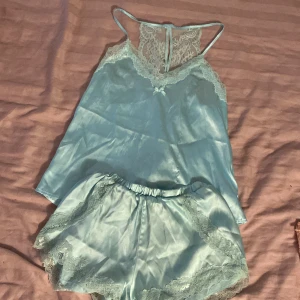 Ljusblå satin linnetopp från Censored - Supersöt ljusblått pyjamasset från Censored, med tunna axelband och spetsdetaljer både fram och bak. Toppen har en liten rosett framtill och vacker spets längs halsringningen och ryggen. Toppen är i storlek XS och shortsen är S.