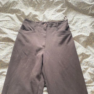 Svarta högmidjade straight kostymbyxor - Säljer ett par mörkbruna högmidjade kostymbyxor med straight ben och klassisk passform. Byxorna har hög midja, bälteshällor och två fickor fram. Perfekta för en clean och stilren look. Materialet känns mjukt och har en lätt stretch.