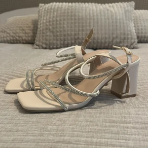 Beiga sandaler med strass och klack - Snygga beiga sandaler med fyrkantig tå och bred klack. Remmarna är dekorerade med glittrande strass och har en elegant design med spänne vid ankeln. 