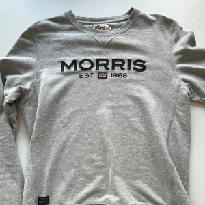Grå sweatshirt från Morris - Snygg grå sweatshirt från Morris med broderad logga och brittisk flagga framtill. Tröjan har rund halsringning och långa ärmar, perfekt för en chill och stilren look. Tillverkad i mjuk bomull för extra komfort.