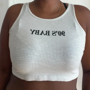 Vit ribbad croppad linnetopp - Säljer en vit ribbad croppad linnetopp med texten '90's BABY' tryckt i svart framtill. Toppen har ärmlös design och rund halsringning, perfekt för en avslappnad och trendig look.