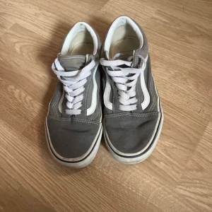 Gråa Vans Old Skool sneakers - Klassiska Vans Old Skool sneakers i grått med vit sula och vit sidostripes. Skorna har snörning och den ikoniska gummisulan med rutmönster. Ovandelen är i canvas och mocka för en snygg och tidlös look.