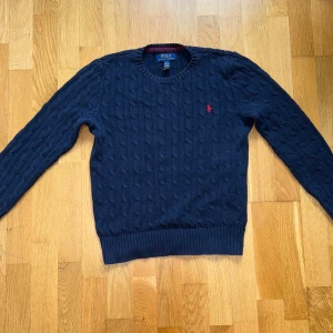 Mörkblå stickad tröja Polo Ralph Lauren - Säljer en klassisk mörkblå stickad tröja från Polo Ralph Lauren med rund hals och det ikoniska röda logobroderiet på bröstet. Tröjan har kabelstickat mönster och långa ärmar, perfekt för lager-på-lager under kyliga dagar. Den är i storlek 10-12 alltså medium i barn storlek. Priset är absolut inte hugget i sten och kom gärna med förslag! Inga förslag är dumma!