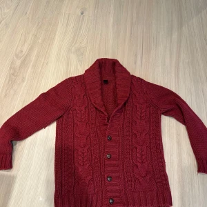 Vinröd stickad kofta med knappar - Säljer en vinröd stickad kofta med grovt kabelstickat mönster framtill och bred krage. Koftan har långa ärmar och stängs med fem knappar. Perfekt för kyliga dagar och enkel att styla över en t-shirt eller skjorta.