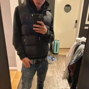 Ralph lauren väst  - Snygg svart dunväst från Ralph Lauren med dragkedja och broderad röd logga på bröstet. Västen har praktiska fickor med dragkedjor och är perfekt för kyligare dagar.