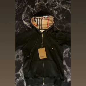 Svart Burberry hoodie med dragkedja - Svart hoodie från Burberry i storlek M med klassiskt rutigt foder i huvan. Tröjan har dragkedja framtill, snörning vid huvan och ribbade muddar. Perfekt för dig som vill ha en stilren och lyxig look med ikoniska Burberry-detaljer.