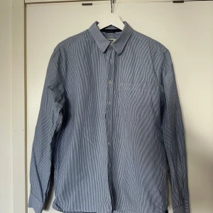 Randig blå skjorta från Dobber - Snygg blå- och vitrandig skjorta från Dobber med klassisk krage och bröstficka. Skjortan har långa ärmar och knäppning framtill. Perfekt för dig som gillar en stilren och tidlös look.