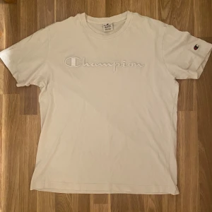 Vit Champion t-shirt med broderi - Snygg vit t-shirt från Champion med broderad logga över bröstet och liten Champion-märke på ärmen. Klassisk rund hals och korta ärmar. Perfekt för dig som gillar enkel och stilren streetwear.
