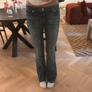 Blå bootcut jeans med färg detaljer - Snygga blå jeans med bootcut passform och låg midja. Jeansen har guldiga färg detaljer, råa kanter vid benslut och coola fickor med silverknappar bak. Materialet är klassisk denim och tvätten har en lätt urtvättad look. Storlek 34. 
