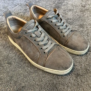 Grå mockasneakers med röd sula - Snygga grå sneakers i mocka från Christian Louboutin med klassisk röd sula. Skorna har rund tå, snörning och vita kanter runt sulan. Perfekta för dig som vill ha en stilren men ändå unik look.