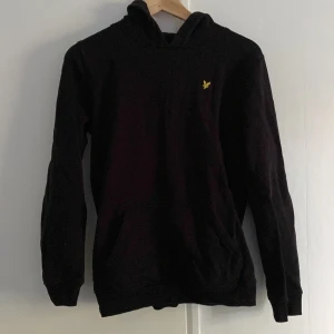 Svart hoodie från Lyle & Scott Junior - Svart hoodie från Lyle & Scott Junior med klassisk gul broderad logga på bröstet. Tröjan har huva, känguruficka och är långärmad. Skriv vid intresse