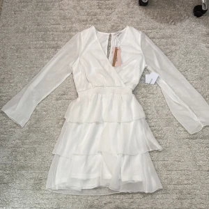 Vit omlottklänning med volanger Bubbleroom - Supersöt vit kortklänning från Bubbleroom med omlottliv, långa transparenta ärmar och flera lager volanger i kjolen. Perfekt för dig som gillar romantisk stil och vill ha något extra till vår eller sommar. Klänningen har v-ringning och markerad midja.