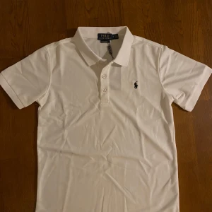 Vit polo t-shirt  - Vit polo t-shirt  i bomull med klassisk krage och tre knappar framtill. Custom slim fit-modell med korta ärmar och den ikoniska svarta broderad på bröstet. Perfekt för en clean och stilren look.