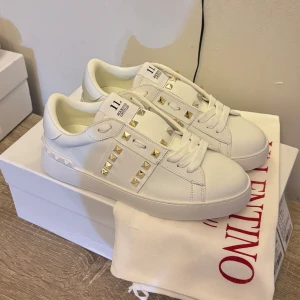 Valentino Rockstud sneakers i vitt skinn - Säljer ett par vita Valentino Rockstud sneakers med coola guldiga nitar längs sidorna och på hälen. Skorna har snörning, platt sula och rund tå. Märket syns på plösen och innersulan. Perfekt för dig som vill sticka ut med lyxig streetstyle.Allt originellt finns kvar