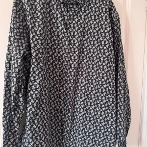 Svartvit blommig skjorta från Riley - Snygg svart skjorta från Riley i super slim fit med vitt blommönster över hela tyget. Skjortan är tillverkad i 100% bomull och har långa ärmar samt klassisk krage. Perfekt för dig som gillar att sticka ut med stilrena detaljer.