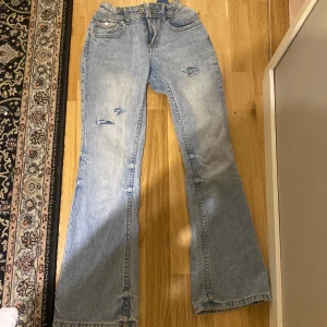 Ljusblå bootcut jeans med slitningar - Säljer ett par ljusblå bootcut jeans med snygga slitningar på låren. Jeansen har låg midja och bandet inuti jeansen går att justera. Längden är 158 cm och storleken är 11-12 år från h&m. Perfekta för en höstdag med en mysig tröja!.